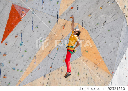 Sportsman climber hanging with one hand on an artificial climbing wall outdoors. Sportsman climber hanging with one hand on an artificial climbing wall outdoors. 92521510