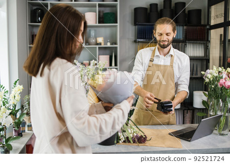 Bearded florist man holding card reader machine at counter with customer paying with credit card. 92521724