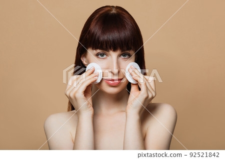 Woman with red hair and bare shoulders posing in studio with cotton pads in hands. 92521842