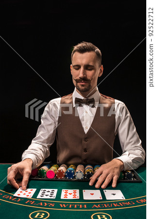 Portrait of a croupier is holding playing cards, gambling chips on table. Black background 92522712