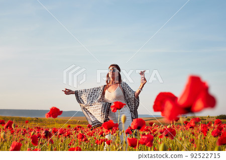 A young woman poses in a field with red wild poppies at sunset. She is holding a glass of strawberries. Front view. A young woman poses in a field with red wild poppies at sunset. She is holding a glass of strawberries. Front view. 92522715