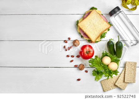 Lunch boxes with sandwich and fresh vegetables, bottle of water, nuts and eggs on white wooden background. Top view with copy space 92522748