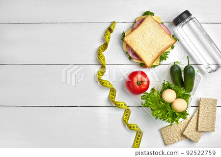Lunch boxes with sandwich and fresh vegetables, bottle of water and eggs on white wooden background. Top view with copy space Lunch boxes with sandwich and fresh vegetables, bottle of water and eggs on white wooden background. Top view with copy space 92522759
