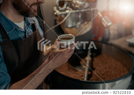 Close up of male barista hands holding cup of coffee on background of roasting coffee machine Close up of male barista hands holding cup of coffee on background of roasting coffee machine 92522810