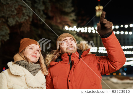 Horizontal shot of young couple walking on winter evening, man showing something to his girlfriend Horizontal shot of young couple walking on winter evening, man showing something to his girlfriend 92523910