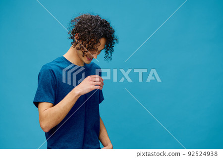 Smiling joyful cute curly tanned Caucasian guy in basic t-shirt posing isolated on over blue background. Lifestyle and Emotions concept. Good offer with copy space for ad Smiling joyful cute curly tanned Caucasian guy in basic t-shirt posing isolated on over blue background. Lifestyle and Emotions concept. Good offer with copy space for ad 92524938