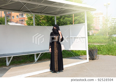 A Muslim woman in black national clothes stands alone at a bus stop in the city A Muslim woman in black national clothes stands alone at a bus stop in the city 92528504