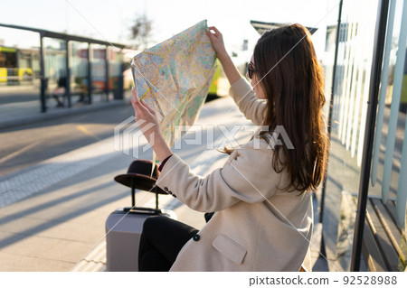 Young woman sitting at bus stop and holding city map. Girl looks at the paper map and search for the location she needs. Travel concept. Young woman sitting at bus stop and holding city map. Girl looks at the paper map and search for the location she needs. Travel concept. 92528988