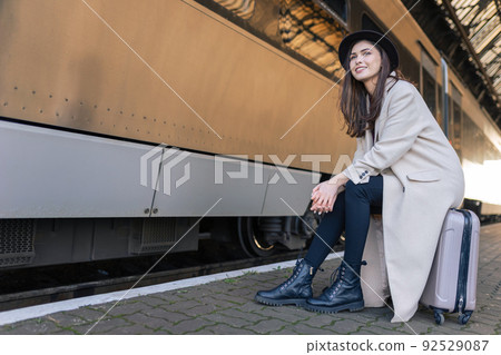 Girl sitting on the suitcase waiting for train depart. Stylish traveler on the platform of the railway station Girl sitting on the suitcase waiting for train depart. Stylish traveler on the platform of the railway station 92529087