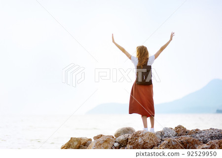 Charming young tourist woman on the Mediterranean coast. Attractive red-haired girl hiking by the sea shore. 92529950