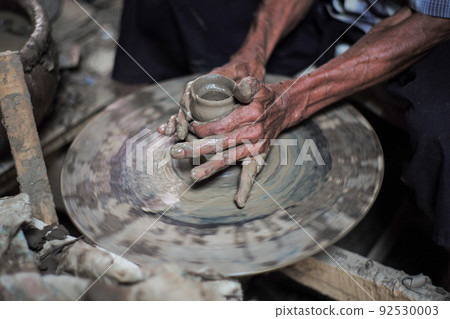 Selective focused on the dirty wrinkled skin hands of old man molding the clay work on the spinning wheel for making the traditional jar 92530003