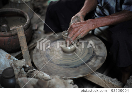 Selective focused on the dirty wrinkled skin hands of old man molding the clay work on the spinning wheel for making the traditional jar 92530004