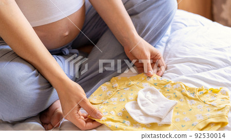 Pregnancy baby clothes. Beautiful pregnant mother with yellow baby clothes. Pregnancy woman hugging belly and packing maternity hospital bag. Concept maternity, pregnancy, childbirth. Pregnancy baby clothes. Beautiful pregnant mother with yellow baby clothes. Pregnancy woman hugging belly and packing maternity hospital bag. Concept maternity, pregnancy, childbirth. 92530014