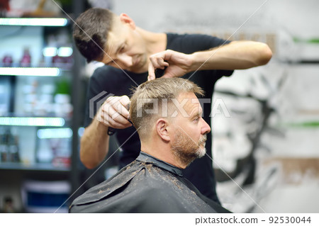Hairdresser is cutting hair of handsome bearded mature man in salon. Stylist making hairstyle with electric shaver for person in barbershop. Services of a professional stylist. Fashion haircut Hairdresser is cutting hair of handsome bearded mature man in salon. Stylist making hairstyle with electric shaver for person in barbershop. Services of a professional stylist. Fashion haircut 92530044