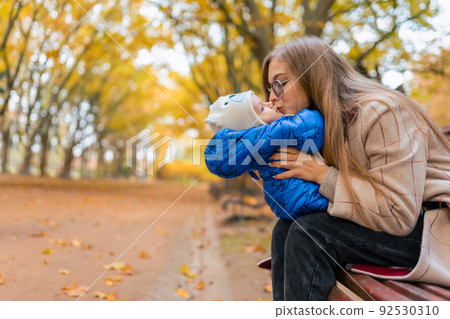 Mother with baby in hands on park bench. Mom kisses infant 92530310