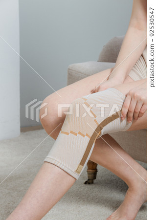 Knee support brace on a woman leg. girl in an orthosis in the interior of the house. Orthopedic anatomic braces for knee fixation, injuries and pain. Knee Joint Bandage Sleeve. Elastic Sports. Knee support brace on a woman leg. girl in an orthosis in the interior of the house. Orthopedic anatomic braces for knee fixation, injuries and pain. Knee Joint Bandage Sleeve. Elastic Sports. 92530547