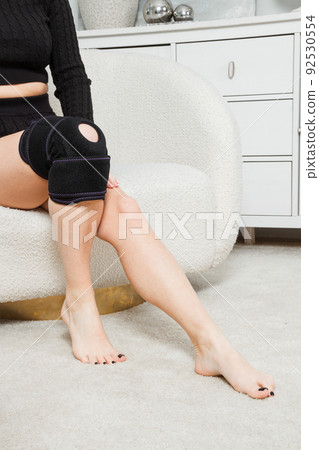 Black knee support brace on a woman leg. girl in an orthosis in the interior of the house. Orthopedic anatomic braces for knee fixation, injuries and pain. Knee Joint Bandage Sleeve. Elastic Sports. Black knee support brace on a woman leg. girl in an orthosis in the interior of the house. Orthopedic anatomic braces for knee fixation, injuries and pain. Knee Joint Bandage Sleeve. Elastic Sports. 92530554