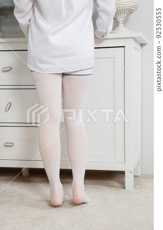 White hosiery. Beautiful long female legs in stockings. Girl putting on stockings at home in a white room. Varicose veins prevention. Woman body in underwear. 92530555