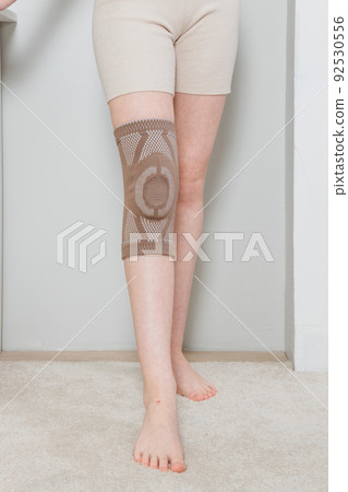 Knee support brace on a woman leg. girl in an orthosis in the interior of the house. Orthopedic anatomic braces for knee fixation, injuries and pain. Knee Joint Bandage Sleeve. Elastic Sports. Knee support brace on a woman leg. girl in an orthosis in the interior of the house. Orthopedic anatomic braces for knee fixation, injuries and pain. Knee Joint Bandage Sleeve. Elastic Sports. 92530556