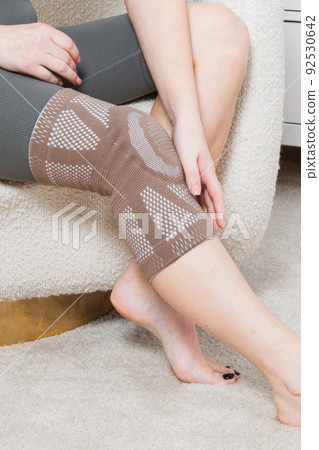 Knee support brace on a woman leg. girl in an orthosis in the interior of the house. Orthopedic anatomic braces for knee fixation, injuries and pain. Knee Joint Bandage Sleeve. Elastic Sports. Knee support brace on a woman leg. girl in an orthosis in the interior of the house. Orthopedic anatomic braces for knee fixation, injuries and pain. Knee Joint Bandage Sleeve. Elastic Sports. 92530642
