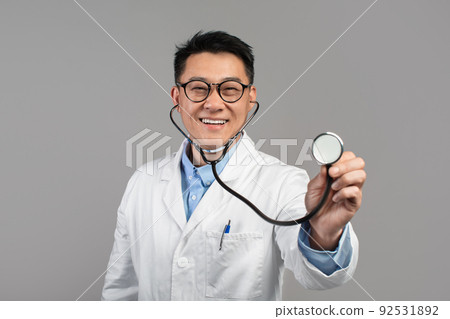 Cheerful confident mature japanese doctor in white coat and glasses listens camera with stethoscope Cheerful confident mature japanese doctor in white coat and glasses listens camera with stethoscope 92531892