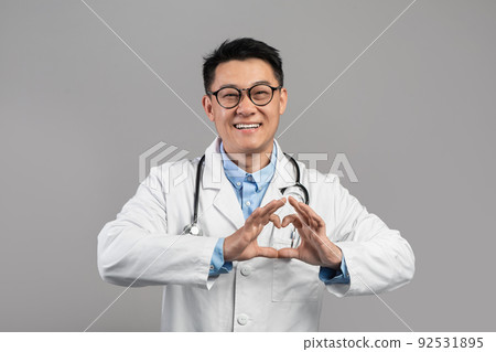 Cheerful middle aged japanese male doctor in white coat and glasses shows heart with hands isolated Cheerful middle aged japanese male doctor in white coat and glasses shows heart with hands isolated 92531895