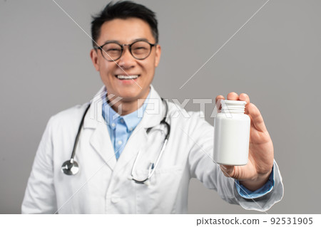 Smiling mature asian man doctor in coat, glasses, shows jar of medicines isolated on gray background, close up Smiling mature asian man doctor in coat, glasses, shows jar of medicines isolated on gray background, close up 92531905