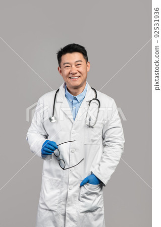 Smiling confident adult chinese man therapist in white coat, protective gloves hold glasses, look at camera Smiling confident adult chinese man therapist in white coat, protective gloves hold glasses, look at camera 92531936