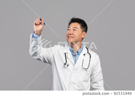 Smiling adult asian guy doctor in white coat with stethoscope writes on virtual blackboard with blank space 92531940