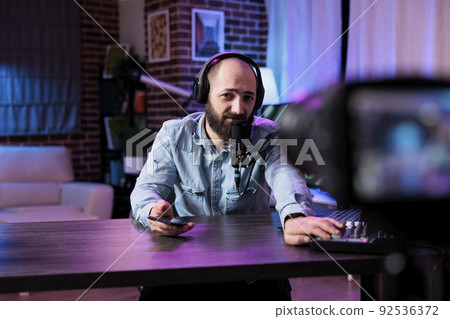 Male influencer filming vlog with podcast equipment in studio, recording online video and hosting talk show at station. Content creator using live broadcast on entertainment channel. Male influencer filming vlog with podcast equipment in studio, recording online video and hosting talk show at station. Content creator using live broadcast on entertainment channel. 92536372