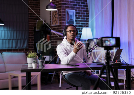 African american blogger recording live podcast vlog on camera, filming livestream in studio with neon lights. Social media influencer vlogging internet video as channel content. African american blogger recording live podcast vlog on camera, filming livestream in studio with neon lights. Social media influencer vlogging internet video as channel content. 92536380