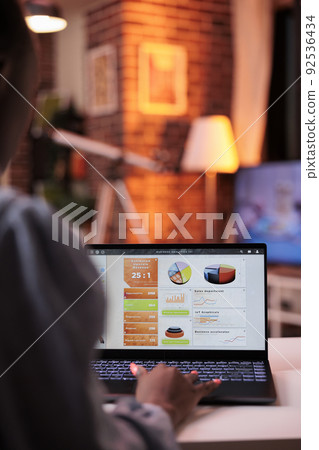 Company research results diagrams and pie charts on laptop screen. African american female remote corporate worker analyzing business statistics in home office in evening, focus on computer 92536434