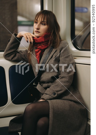 young caucasian woman with brown hair wearing elegant classy coat and a scarf sitting alone in subway carriage and distantly looking out window. Image with selective focus, toning and noise effect 92540995