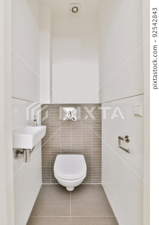 A modern toilet is installed on a beige wall under a button and a small ceramic sink in a light restroom at home 92542843