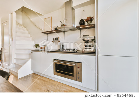 A fragment of the interior of a light home kitchen in a minimalist style with a sink, stove, extractor hood and with white furniture next to the stairs A fragment of the interior of a light home kitchen in a minimalist style with a sink, stove, extractor hood and with white furniture next to the stairs 92544683