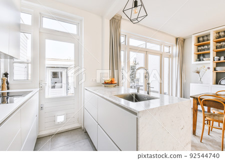 Interior of spacious light dining room with elegant chandelier located near kitchen with comfortable furniture and appliances 92544740