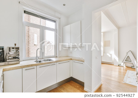 Fragment of interior of light narrow home kitchen with minimalist style white furniture with sink and stove Fragment of interior of light narrow home kitchen with minimalist style white furniture with sink and stove 92544927