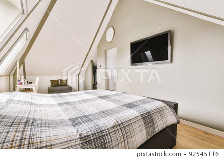 Comfortable bed placed near white wall in modern minimalist style mansard bedroom with windows and tv Comfortable bed placed near white wall in modern minimalist style mansard bedroom with windows and tv 92545116