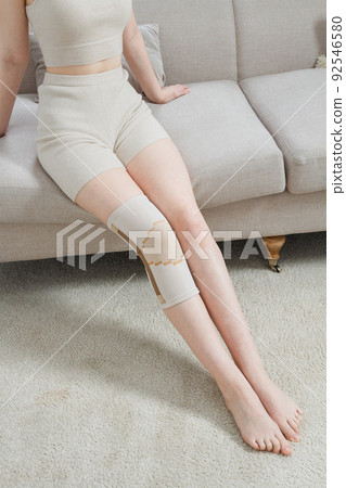 Knee support brace on a woman leg. girl in an orthosis in the interior of the house. Orthopedic anatomic braces for knee fixation, injuries and pain. Knee Joint Bandage Sleeve. Elastic Sports. Knee support brace on a woman leg. girl in an orthosis in the interior of the house. Orthopedic anatomic braces for knee fixation, injuries and pain. Knee Joint Bandage Sleeve. Elastic Sports. 92546580