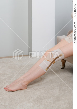 Knee support brace on a woman leg. girl in an orthosis in the interior of the house. Orthopedic anatomic braces for knee fixation, injuries and pain. Knee Joint Bandage Sleeve. Elastic Sports. 92546597