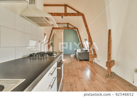 Spacious attic room of apartment with dining table and chairs near open kitchen under roof with beams Spacious attic room of apartment with dining table and chairs near open kitchen under roof with beams 92546652