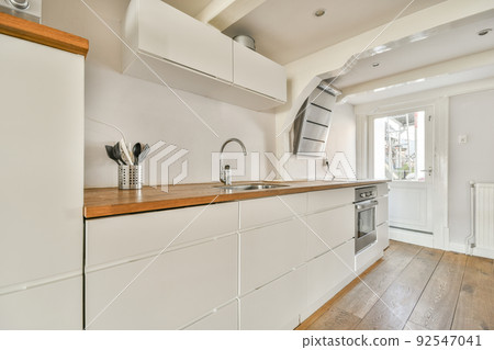 Contemporary cupboards with appliances and tables with kitchenware located in light kitchen near dining room in stylish apartment 92547041