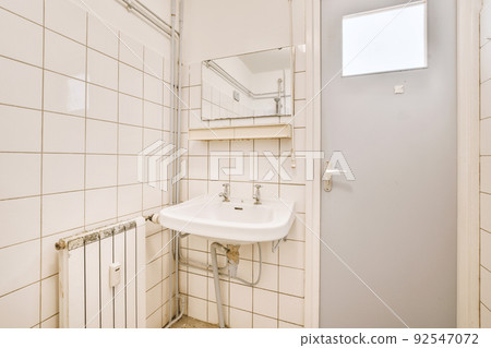 Cupboard with sink and mirror attached to tiled wall near door and drying rack in modern restroom Cupboard with sink and mirror attached to tiled wall near door and drying rack in modern restroom 92547072