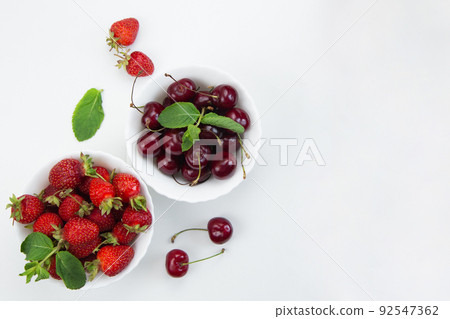 Two white plates with cherries and strawberries with mint petals stand on a white background with space for text. High quality photo Two white plates with cherries and strawberries with mint petals stand on a white background with space for text. High quality photo 92547362