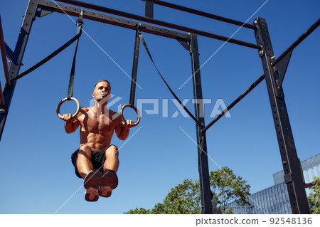 Handsome man on sport ground. The sportsman the guy, carries out difficult exercise, sports gymnastics. Muscular man doing workout with rings. The concept of a modern young man 92548136