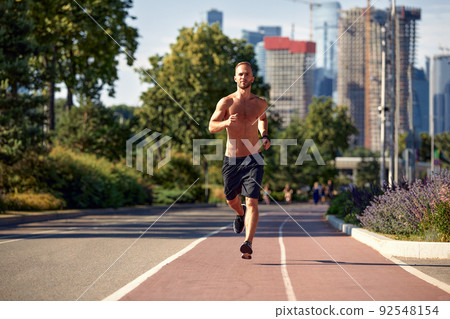 caucasian man jogging on the running track - Blonde male athlete in stadium training run in sunny spring or summer day - real people healthy lifestyle concept 92548154