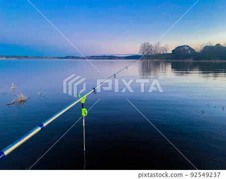 A silver fishing rod on a green holder stands abandoned in the lake against the background of blue water, the morning sky beginning to lighten and a small grove of trees on the other side. High 92549237