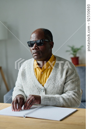 African blind man in dark glasses sitting at table and reading book in braille language 92550693