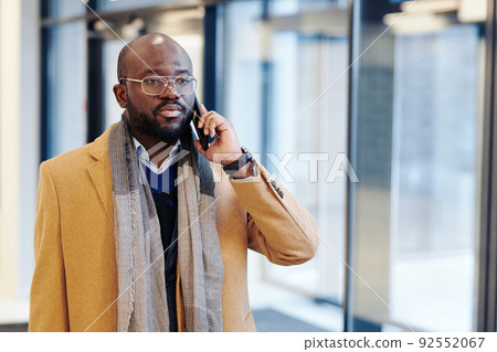 African businessman in stylish coat talking on mobile phone while walking along the office corridor after work 92552067