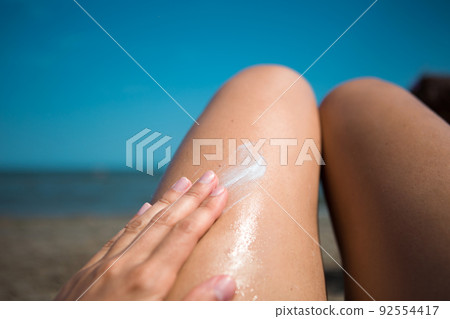 A woman on the beach smears her feet with sunscreen lotion from an orange tube. Skin care, UV protection, sunscreen. Beach holidays 92554417